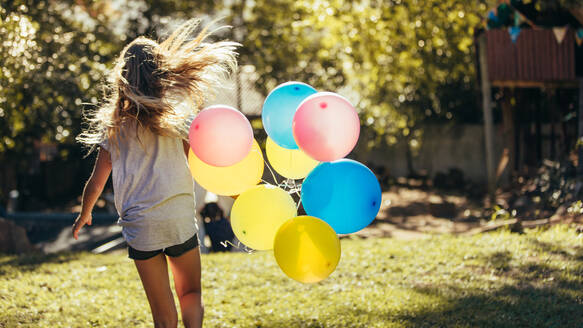 Rückansicht eines kleinen Mädchens, das mit bunten Luftballons im Hinterhof davonläuft. Mädchen hat Spaß im Freien mit Luftballons. - JLPSF20638
