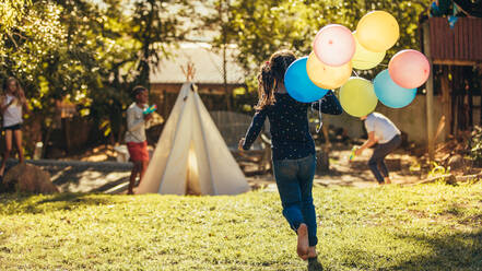 Kleines Mädchen mit bunten Luftballons läuft auf spielende Kinder im Garten zu. Kinder haben Spaß im Freien. - JLPSF20639