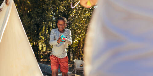 Little african boy playing squirt gun fight with friend in backyard. Kids enjoying playing water gun fight outdoors. - JLPSF20642