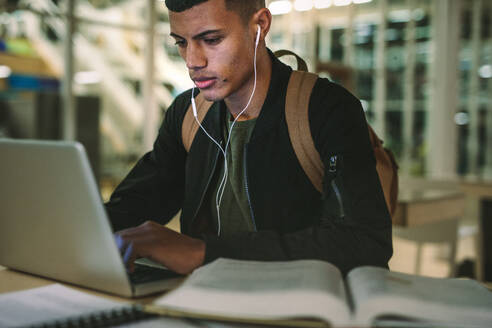 Junger männlicher Student, der eine Schulaufgabe in der Bibliothek vorbereitet. Mann mit Kopfhörern und Tasche arbeitet am Laptop in der Universitätsbibliothek. - JLPSF20654