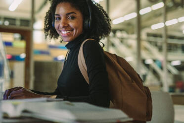 Glückliche afroamerikanische Studentin mit Laptop und Büchern am Tisch sitzend und lächelnd in die Kamera schauend. Studentin studiert in der Bibliothek. - JLPSF20656