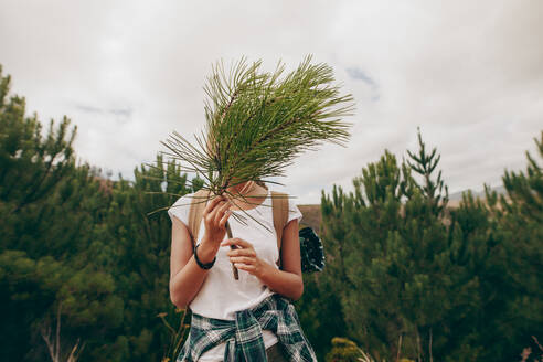 Frau, die ihr Gesicht mit einem kleinen Zweig bedeckt, mit Bäumen im Hintergrund. Frau, die einen Rucksack trägt und durch einen Wald reist. - JLPSF20661