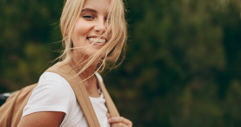 Close up of woman on a holiday walking in a park wearing a bag. Portrait of a smiling woman with brown hair flying over her face. - JLPSF20664