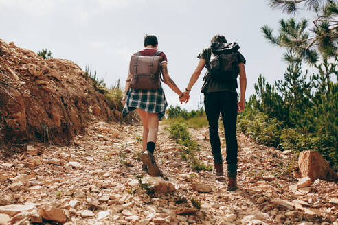 Rückansicht eines Mannes und einer Frau, die zusammen durch unwegsames Gelände wandern und sich an den Händen halten. Ein Paar, das im Urlaub Rucksäcke trägt und auf einem Hügel wandert. - JLPSF20673
