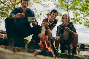 Zwei Frauen rösten Marshmallows am Lagerfeuer mit Stöcken, während ein Mann eine Kaffeetasse hält. Lächelnde Freunde rösten Essen am Lagerfeuer in der Natur. - JLPSF20684