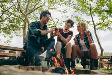 Smiling friends on holiday toasting food on bonfire. Friends making a sandwich with toasted marshmallows sitting outdoors in the countryside. - JLPSF20685