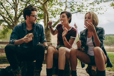 Tourist friends on vacation eating toasted snacks sitting outdoors in the country side. Man with two female friends camping in the country side eating snacks. - JLPSF20686