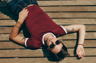 Top view of a woman on a holiday sunbathing lying on a wooden floor. Woman wearing sunglasses lying on a floating dock on a sunny day. - JLPSF20692