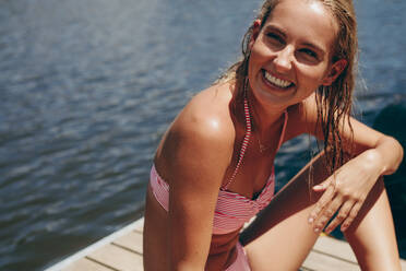 Frau im Bikini genießt ihren Urlaub an einem See. Frau im Badeanzug sitzt auf einem schwimmenden Steg an einem See. - JLPSF20695