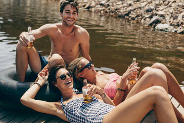 Mann mit zwei Freundinnen, die auf einem schwimmenden Steg in einem See sitzen und Bier trinken. Freunde, die sich an einem See amüsieren und sich mit Getränken in der Hand entspannen. - JLPSF20696