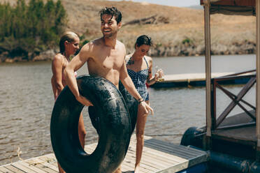 Friends standing on floating dock after swimming in the lake. Man standing near a lake with an inflatable rubber tube with his female friends drinking beer. - JLPSF20703