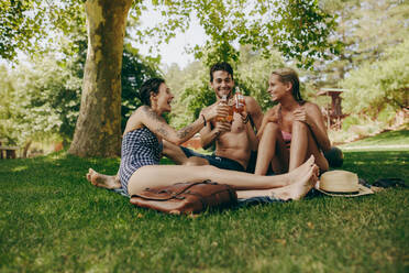 Zwei Frauen im Bikini stoßen mit einem männlichen Freund in einem Park auf ihre Getränke an. Freunde genießen und entspannen im Park mit Getränken in der Hand. - JLPSF20704