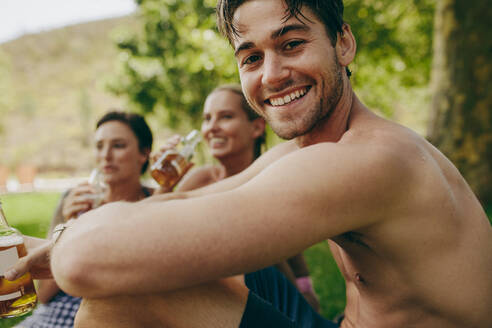 Freunde genießen ihren Urlaub in einem Park mit Getränken. Ein Mann und zwei Freundinnen sitzen im Freien und trinken Bier. - JLPSF20707