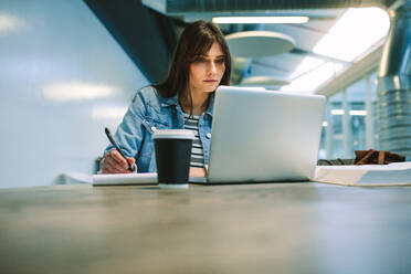 Smart female student at university campus looking at laptop computer and writing notes. Woman student studying at college canteen. - JLPSF20711