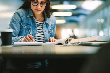 Studentin schreibt Notizen aus einem Lehrbuch in einen Notizblock. Studentin studiert in der Universitätsbibliothek. - JLPSF20717