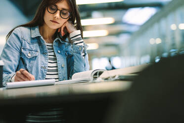 Junge Frau sitzt in der Bibliothek eines Colleges und lernt. Mädchen schreibt Notizen auf dem College-Campus. - JLPSF20719