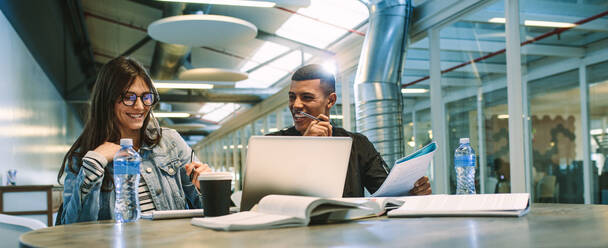 Smiling man and woman sitting at table with books and laptop. Classmates working together on college assignments. - JLPSF20728