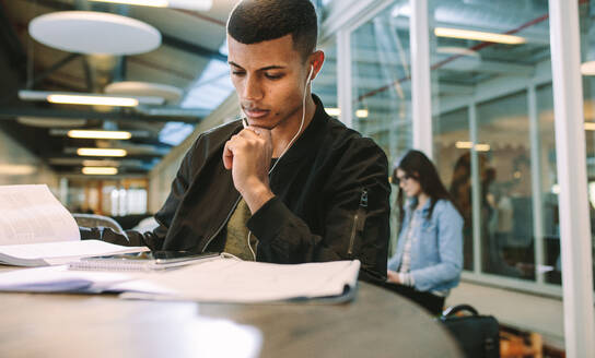 Mann mit Kopfhörern, der in der Universitätsbibliothek sitzt und auf ein digitales Tablet schaut. Männlicher Student, der auf dem College-Campus studiert und ein digitales Tablet benutzt. - JLPSF20732