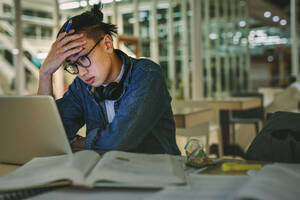 Männlicher Student sitzt in der Bibliothek und schaut auf den Laptop mit der Hand auf der Stirn. Depressiver Student sitzt in der Bibliothek mit dem Laptop im College. - JLPSF20751