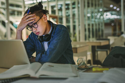 Männlicher Student sitzt in der Bibliothek und schaut auf den Laptop mit der Hand auf der Stirn. Depressiver Student sitzt in der Bibliothek mit dem Laptop im College. - JLPSF20751