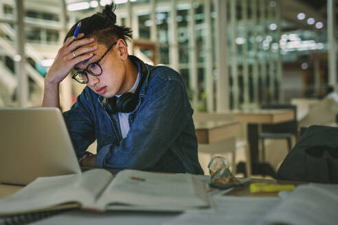 Male student sitting at library looking at laptop with hand on forehead. Depressed student sitting in library with laptop in college. - JLPSF20751