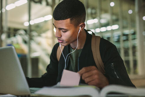 Männlicher Student sitzt und macht Notizen mit Laptop auf dem Tisch und bereitet Klassenarbeiten vor. Junger Mann studiert in der College-Bibliothek. - JLPSF20754