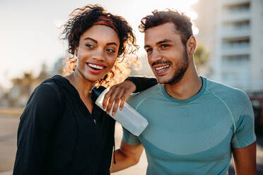 Couple in fitness wear relaxing outdoors in the morning. Smiling couple standing together taking a break after work out. - JLPSF20759