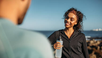 Lächelnde Frau, die sich nach dem Training entspannt und Wasser trinkt. Frau im Gespräch mit ihrer Freundin nach dem Training im Freien. - JLPSF20760