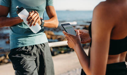 Athletic couple standing outdoors together taking a break after running. Man looking at his wrist watch holding a water bottle while his female friend is looking at mobile phone. - JLPSF20762