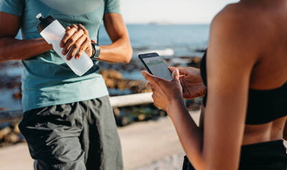 Ein sportliches Paar steht im Freien und macht eine Pause nach dem Laufen. Der Mann schaut auf seine Armbanduhr und hält eine Wasserflasche, während seine Freundin auf ihr Handy schaut. - JLPSF20762