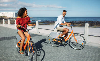 Ein glücklicher Mann und eine Frau fahren an einem sonnigen Tag mit dem Meer im Hintergrund Fahrrad. Ein Touristenpaar fährt mit dem Fahrrad durch die Stadt. - JLPSF20764