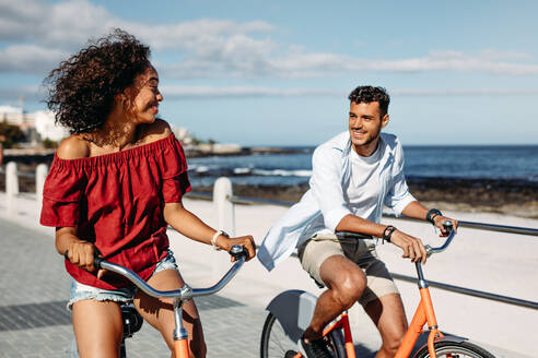 Lächelndes Paar, das sich in der Stadt auf Fahrrädern fortbewegt und einander ansieht. Mann und Frau genießen die gemeinsame Zeit beim Fahrradfahren auf der Straße. - JLPSF20765