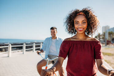 Smiling woman riding a bicycle on the street with her boyfriend following her. Happy couple riding bicycles on a sunny day with sea in the background. - JLPSF20771