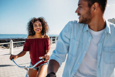 Smiling woman riding a bicycle on a street looking at her boyfriend. Happy man and woman having fun riding bicycles on a sunny day. - JLPSF20773