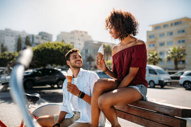 Romantic couple sitting on a bench in street eating cone ice cream looking at each other. Young man and woman having fun moving around the city eating ice cream. - JLPSF20775