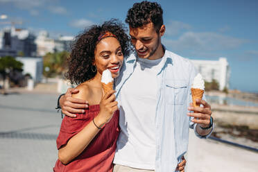 Verliebtes Paar, das auf der Straße spazieren geht und Eis isst. Junger Mann und Frau verbringen Zeit miteinander, indem sie auf der Straße spazieren gehen, sich unterhalten und Eis essen. - JLPSF20777