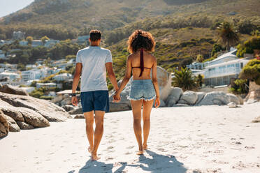 Rear view of a tourist couple walking on beach on a sunny day. Couple walking on the beach together holding hands. - JLPSF20780