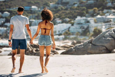 Rückansicht eines Paares, das an einem felsigen Strand spazieren geht und sich an den Händen hält. Paar im Urlaub, das an einem sonnigen Tag zusammen am Strand spazieren geht. - JLPSF20783