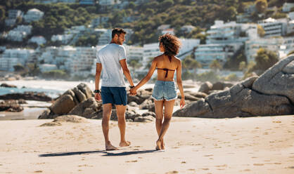 Tourist couple on vacation walking on beach looking at each other. Rear view of a couple walking on a rocky beach hand in hand on a sunny day. - JLPSF20784