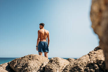 Rückansicht eines Mannes ohne Hemd, der auf einem Felsen am Meer steht. Ein Tourist genießt die landschaftliche Schönheit des Meeres am Strand. - JLPSF20793