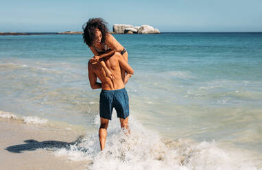 Rear view of a romantic tourist couple having fun playing at the beach. Man walking in sea water lifting his girlfriend on his shoulder. - JLPSF20797