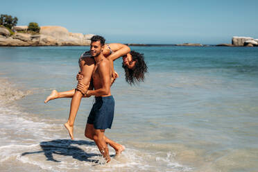 Mann trägt seine Freundin auf der Schulter, die im Meerwasser am Ufer spazieren geht. Paar genießt und hat Spaß am Meer. - JLPSF20798