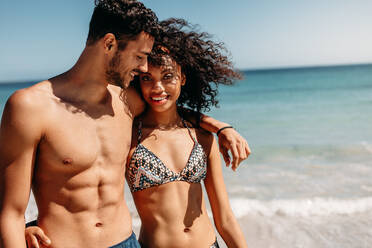 Romantisches Paar, das an einem sonnigen Tag zusammen am Strand spazieren geht. Lächelnder Mann, der mit seiner Freundin am Strand spazieren geht und seinen Arm um ihre Schulter legt. - JLPSF20799