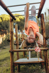 Mädchen hängt kopfüber in einem Park und hält einen herzförmigen Lutscher. Mädchen spielt in einem Park und hängt kopfüber auf einer horizontalen Leiter und genießt einen Lutscher. - JLPSF20812