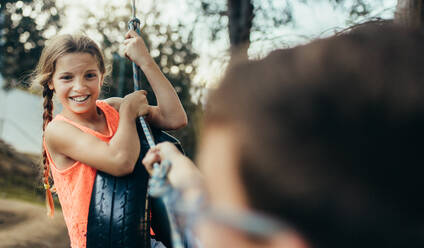 Kinder spielen im Park und haben Spaß beim Schwingen auf einer Reifenschaukel. Mädchen sitzt auf einer Reifenschaukel, die in einem Park hängt, während ein Junge an dem Seil zieht. - JLPSF20814