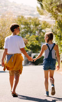 Rückansicht eines Jungen und eines Mädchens, die auf der Straße spazieren gehen und sich anschauen. Kinder, die an einem sonnigen Tag zusammen auf die Straße gehen und sich an den Händen halten. - JLPSF20819