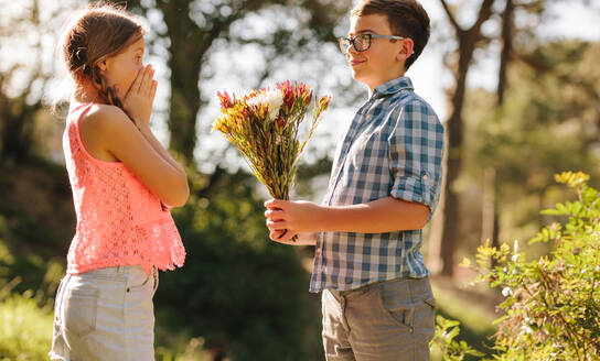 Glücklicher Junge und Mädchen stehen in einem Park, wobei der Junge einen Blumenstrauß überreicht. Aufgeregtes Mädchen drückt seine Überraschung über den Antrag ihres Freundes aus. - JLPSF20826