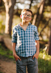 Happy boy standing in park with hands in pocket. Smiling teenage boy in eyeglasses standing in park on a sunny day. - JLPSF20831