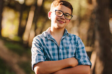Lächelnder Junge mit Brille, der mit verschränkten Armen in einem Park steht. Porträt eines glücklichen Teenagers, der mit verschränkten Armen im Freien steht. - JLPSF20832