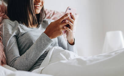 Lächelndes Mädchen sitzt auf dem Bett und überprüft ihr Handy. Mädchen hält ein Handy in beiden Händen und sitzt auf dem Bett. - JLPSF20837